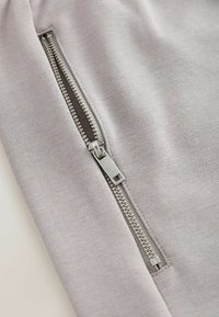 Cerniera in metallo argento parzialmente aperta su tessuto grigio chiaro, che mostra una texture a maglia liscia e cuciture robuste lungo la cucitura della cerniera.