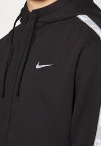Svart zip-up hoodie med dragsko på huvan. Har en liten reflekterande Nike-logga och vita mönstrade detaljer på ärmarna. Slät textur.