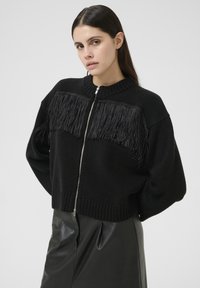 Frau mit langen dunklen Haaren trägt einen schwarzen, mit Reißverschluss versehenen Pullover mit Fransen und einen schwarzen, perforierten Ledrock und steht vor einem schlichten Hintergrund.