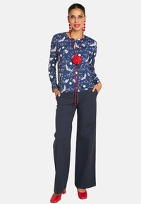 Navyblaue Langarmbluse mit weißem Tiermuster, kombiniert mit lockeren navyblauen Hosen und roten Schuhen. Ergänzt durch eine Halskette mit roter Blume.