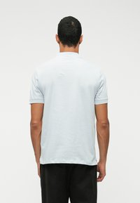 Homem com cabelo curto e escuro, vestindo uma camisa polo de mangas curtas azul claro e calças pretas, de pé, de costas, contra um fundo simples.