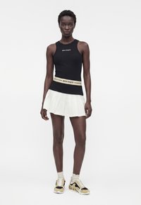 Femme portant un haut noir sans manches avec le texte « palm angels », une jupe plissée blanche, une ceinture assortie et des baskets beige-noir, debout droite.