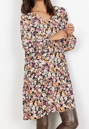 Kvinde iført en langærmet blomsterkjole med pink, orange og hvide blomster kombineret med brune læderbukser.