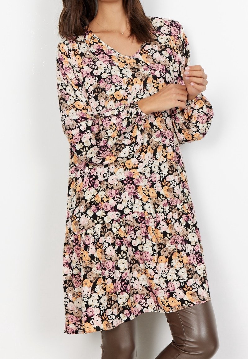 Femme portant une robe à manches longues à motif floral avec des fleurs roses, orange et blanches, associée à un pantalon en cuir marron.