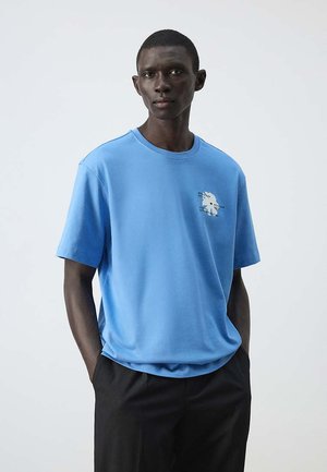 Homme à la peau foncée portant un t-shirt bleu clair à manches courtes avec un petit motif blanc sur la poitrine et un pantalon noir, debout les mains dans les poches.