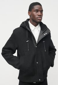 Sort fleecejakke med hætte, forlommer og sølvfarvede trykknapper. Har en glat indvendig foring og justerbare snore.