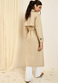 Beige trench takki, jossa on vyötärölle solmittava vyö, kaksirivinen malli ja pitkät hihat. Mallilla on päällään valkoiset leveälahkeiset housut ja ruskeat kengät.
