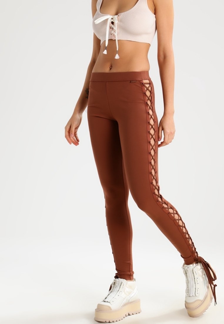 Leggings marrones de corte ajustado con detalles de cordones a lo largo del costado. Combinados con un top corto de color claro y zapatillas blancas de suela gruesa.