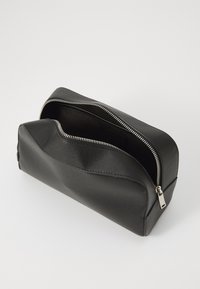 Trousse de maquillage en cuir noir avec une surface texturée, fermeture éclair et forme arrondie. Elle dispose d'un intérieur spacieux avec un seul compartiment.