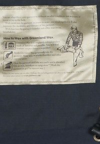 Etichetta di un indumento in tessuto blu navy con istruzioni per l'utilizzo della Greenland Wax; presenta testo in nero e crema, con un'illustrazione di una persona che applica la cera.