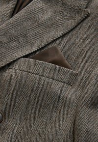 Brązowy blazer w wzór houndstooth z teksturowanym splotem, z solidnym brązowym poszetką wystającą z kieszeni na piersi.