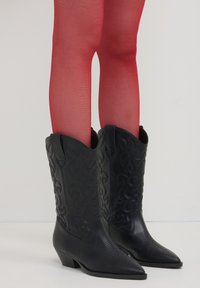 Bottes de cowboy en cuir noir avec des pointes, des coutures décoratives et des talons bas, associées à des collants rouges transparents.