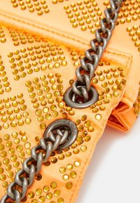 Oranžová látková taška zdobená průhlednými žlutými nýty, s popruhem ze stříbrného kovového řetězu a kruhovým kovovým detailem.