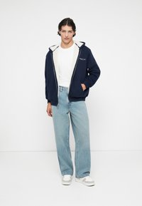 Hoodie bleu marine avec une doublure en fausse fourrure blanche et une fermeture éclair, associé à un jean droit bleu clair et des baskets grises. T-shirt blanc en dessous.