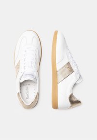 Baskets en cuir blanc avec un détail métallique doré et une semelle en caoutchouc texturée. Dotées de lacets blancs et d'un détail perforé sur la languette.