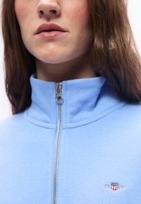 Γυναίκα που φοράει ανοιχτό μπλε τοπ με φερμουάρ και ψηλό γιακά, με μικρό λογότυπο GANT στην αριστερή πλευρά του στήθο υς.