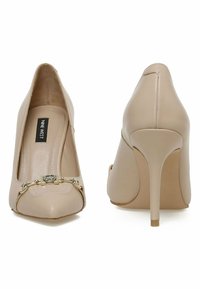 Dei tacchi alti beige con punta affusolata, in pelle liscia, e una catena metallica decorativa sulla parte anteriore. Presentano un alto tacco a stiletto.