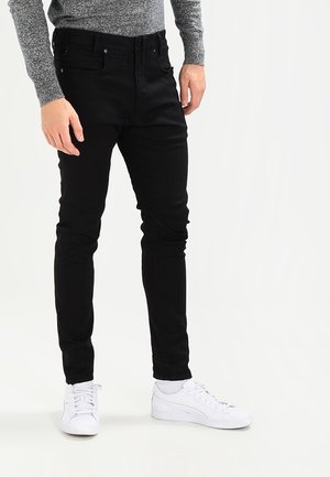 Jeans negros ajustados de mezclilla, con bolsillos frontales y cremallera. Combinados con zapatillas deportivas blancas y un suéter gris.