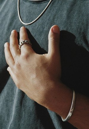 I gioielli d'argento includono una collana sottile, un bracciale liscio e due anelli, uno intrecciato e l'altro semplice, indossati su una mano contro una camicia scura.