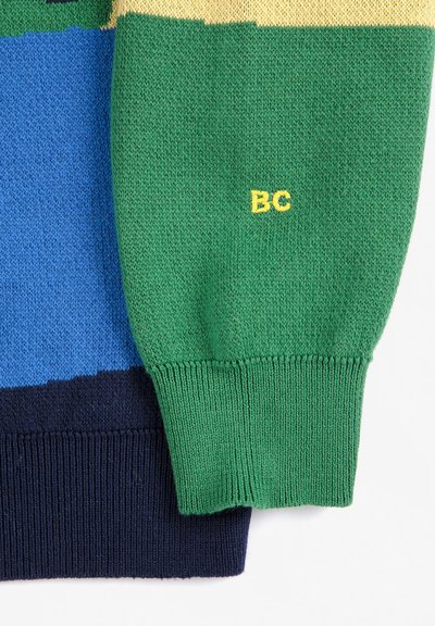 Gros plan d'une manche de pull tricoté avec des blocs de couleur vert, bleu et marine, avec une broderie jaune "BC" sur la partie verte.