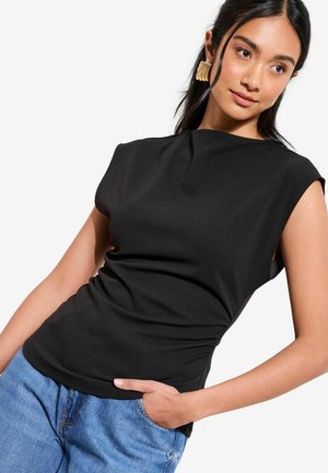 Frau trägt ärmelloses schwarzes Oberteil und blaue Jeans, mit goldenen Statement-Ohrringen, posiert mit einer Hand in der Tasche vor weißem Hintergrund.