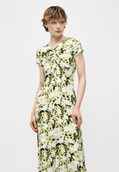 Γυναίκα που φοράει κίτρινο, μαύρο και λευκό φόρεμα με κοντά μανίκια και φλοράλ σχέδιο, με σούρες στον λαιμό σε σχήμα κλειδαρότρυπας, στέκεται μπροστά σε ανοιχτό γκρι φόντο.