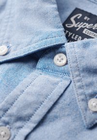 Lichtblauw overhemdd met een gestructureerde stof, puntgekraagd en witte plastic knopen. Bevat een merklabel aan de binnenkant van de kraag.