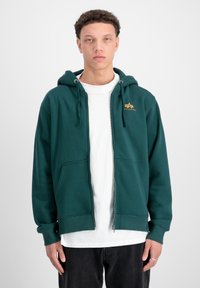 Felpa verde con zip, cappuccio regolabile con cordino, tasche frontali e polsini a costine. Presenta un piccolo logo giallo sul lato sinistro del petto.