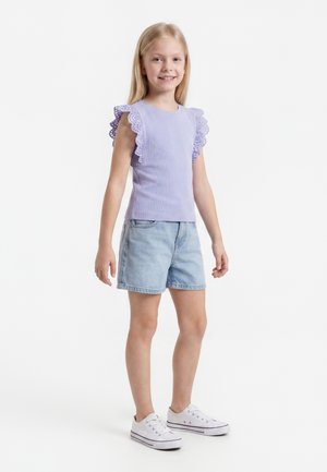 Jong meisje lacht, draagt een lavendelkleurige mouwloze top met gerimpelde mouwen, lichte denimshorts en witte sneakers, staand tegen een witte achtergrond.