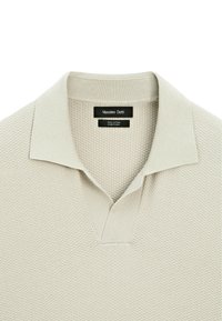 Bomullspolo i ljusbeige med texturerad yta, ribbad krage och v-ringad design. Etiketter visar varumärke och material.