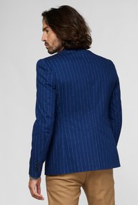 Giacca blu navy a righe con spalle strutturate, dotata di uno spacco singolo e bottoni in tonalità argento, abbinata a pantaloni beige.
