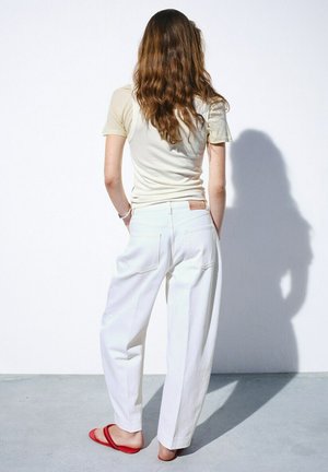 Vrouw met lang bruin haar staat met haar gezicht naar een witte muur gekeerd, draagt een beige top met korte mouwen, witte broek en rode sandalen op een grijze vloer.