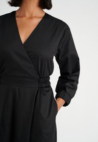Schwarzes Wickelkleid mit V-Ausschnitt, elastischem Taillenumfang und langen Ärmeln mit gerafften Bündchen. Glatter Stoff, schlichtes Design und ohne Muster.