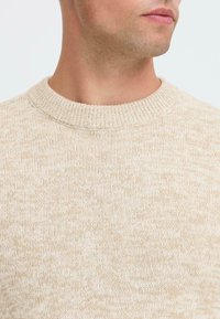 Close-up van de nek en onderkant van het gezicht van een man die een beige gebreide ronde halstrui draagt en naar de zijkant kijkt.