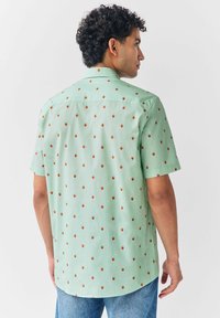 Hombre con cabello rizado vestido con una camisa de manga corta verde claro con pequeños patrones de fresas rojas, combinada con jeans azules, mirando de lado.