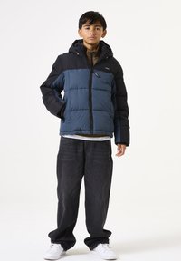 Pufferjack in blauw en zwart met een capuchon, ritssluiting en zijzakken. G gedragen met grijze jeans en witte sneakers tegen een witte achtergrond.