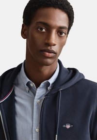 Felpa con cappuccio blu navy con zip, con una camicia a righe blu sotto. Logo ricamato sul petto e dettaglio del cappuccio con coulisse.