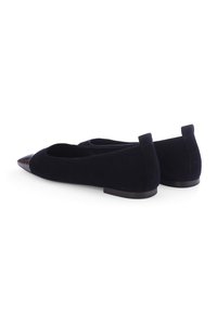 Ein Paar schwarze Wildleder-Ballerinas mit spitzem Zehenbereich und glänzenden schwarzen Vorderkappen, flachen Absätzen und hinteren Absatzlaschen auf einem weißen Hintergrund.