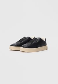 Zwarte leren sneakers met een chunky beige zool, voorzien van een gladde textuur, ronde neus en vetersluiting. Subtiele logo-opdruk aan de zijkant.