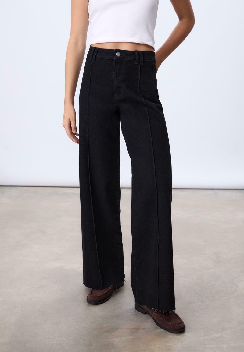 Jean noir à jambes larges avec un ourlet brut, en denim, doté de poches avant et d'une fermeture à bouton. Porté avec des chaussures en suede marron.