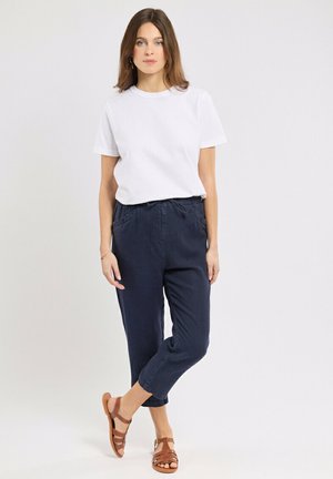 Jeune femme portant un t-shirt blanc à manches courtes, un pantalon bleu marine à cordon et des sandales marron, debout devant un fond clair uni.