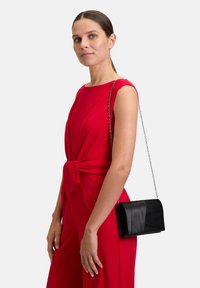 Pochette noire avec un design replié et une surface texturée, dotée d'une bandoulière en chaîne. Portée avec une robe rouge sans manches.