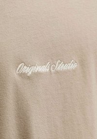 Μπεζ ύφασμα με λευκό κεντημένο κείμενο «Originals Studio» σε καλλιγραφική γραμματοσειρά σε ρούχο.