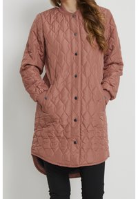 Femme portant un manteau matelassé rose poussière, à mi-length, avec des boutons-pression noirs, des poches latérales et des manches longues, associé à un pantalon noir.