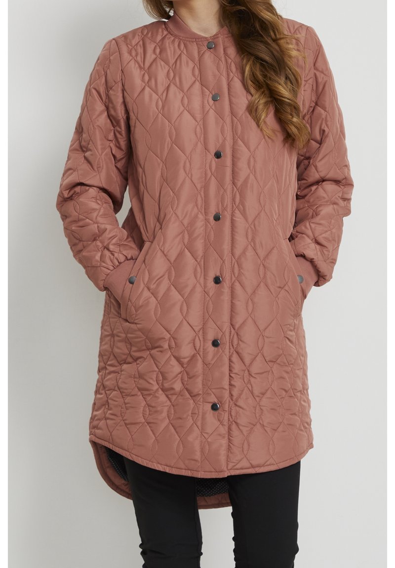 Femme portant un manteau matelassé rose poussière, à mi-length, avec des boutons-pression noirs, des poches latérales et des manches longues, associé à un pantalon noir.