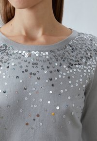 Maglione grigio chiaro con scollatura rotonda, decorato con paillettes argentate e iridescenti sparse sul petto.
