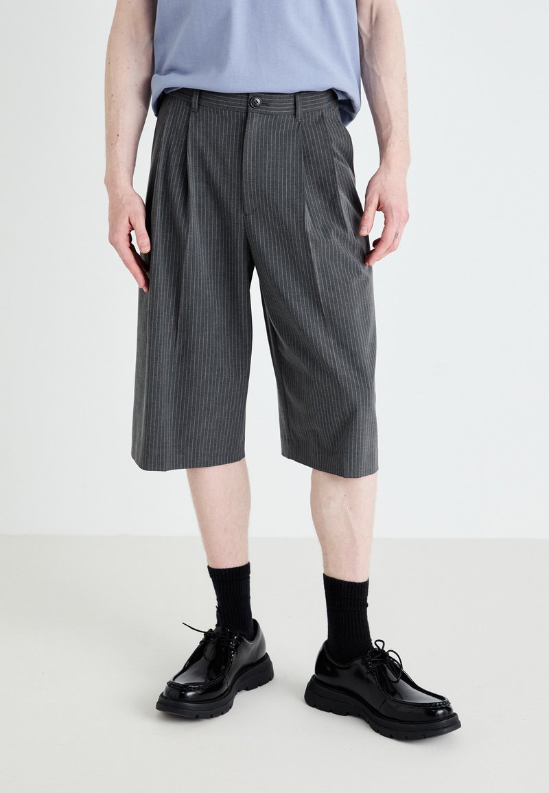Shorts gris à rayures avec des plis, fabriqués dans un tissu léger. Associés à des chaussures noires brillantes et des chaussettes sombres.