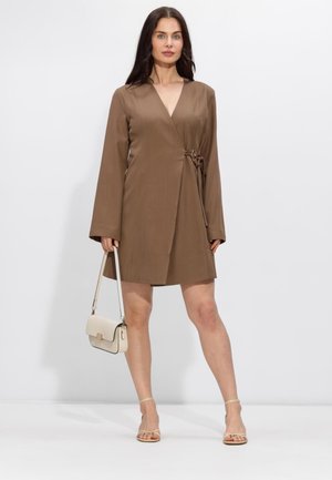 Vrouw met een bruin wikkeljurkje met lange mouwen, een beige handtas in haar handen en beige open tenen hakken, staand tegen een witte achtergrond.