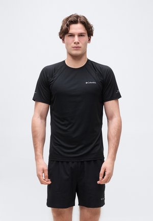 Joven de pie mirando hacia adelante, vistiendo una camisa de manga corta negra de Columbia y pantalones cortos negros a juego, sobre un fondo liso.