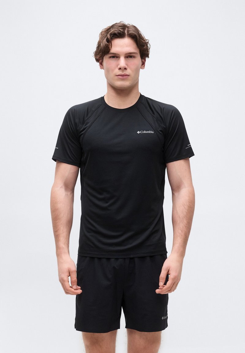 Νέος άνδρας που στέκεται προς τα εμπρός, φοράει μαύρο κοντομάνικο πουκάμισο Columbia και ταιριαστά μαύρα σορτς, μπροστά σε απλό φόντο.